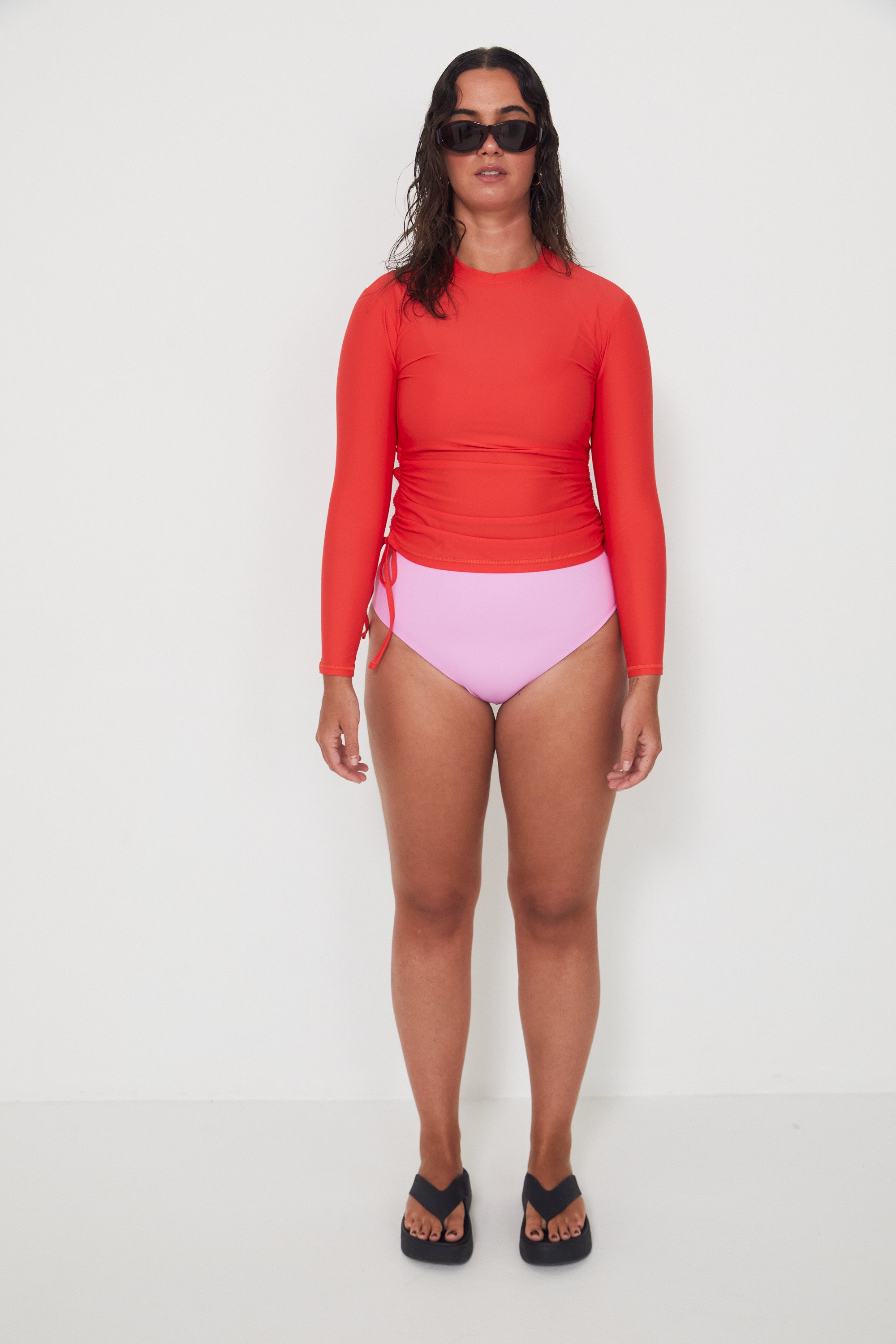 Woman wearing a red long-sleeve  ruched rashguard, salt gypsy swimwear top and pink high-waisted bikini drifter bottoms on a white background, ready for a beach swim