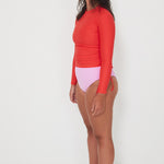Woman wearing a red ruched rashguard long-sleeve swimwear top and pink swimsuit drifter bottom on a white background ready for a swim and surf