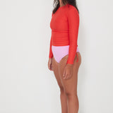 Woman wearing a red ruched rashguard long-sleeve swimwear top and pink swimsuit drifter bottom on a white background ready for a swim and surf