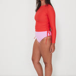 Woman wearing a red ruched rashguard long-sleeve swim top and pink high-waisted drifter bikini bottom on a white background ready for a surf and swim
