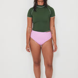 Woman wearing a green olive low rider rashguard and pink drifter swim bottoms with white trim on a plain background