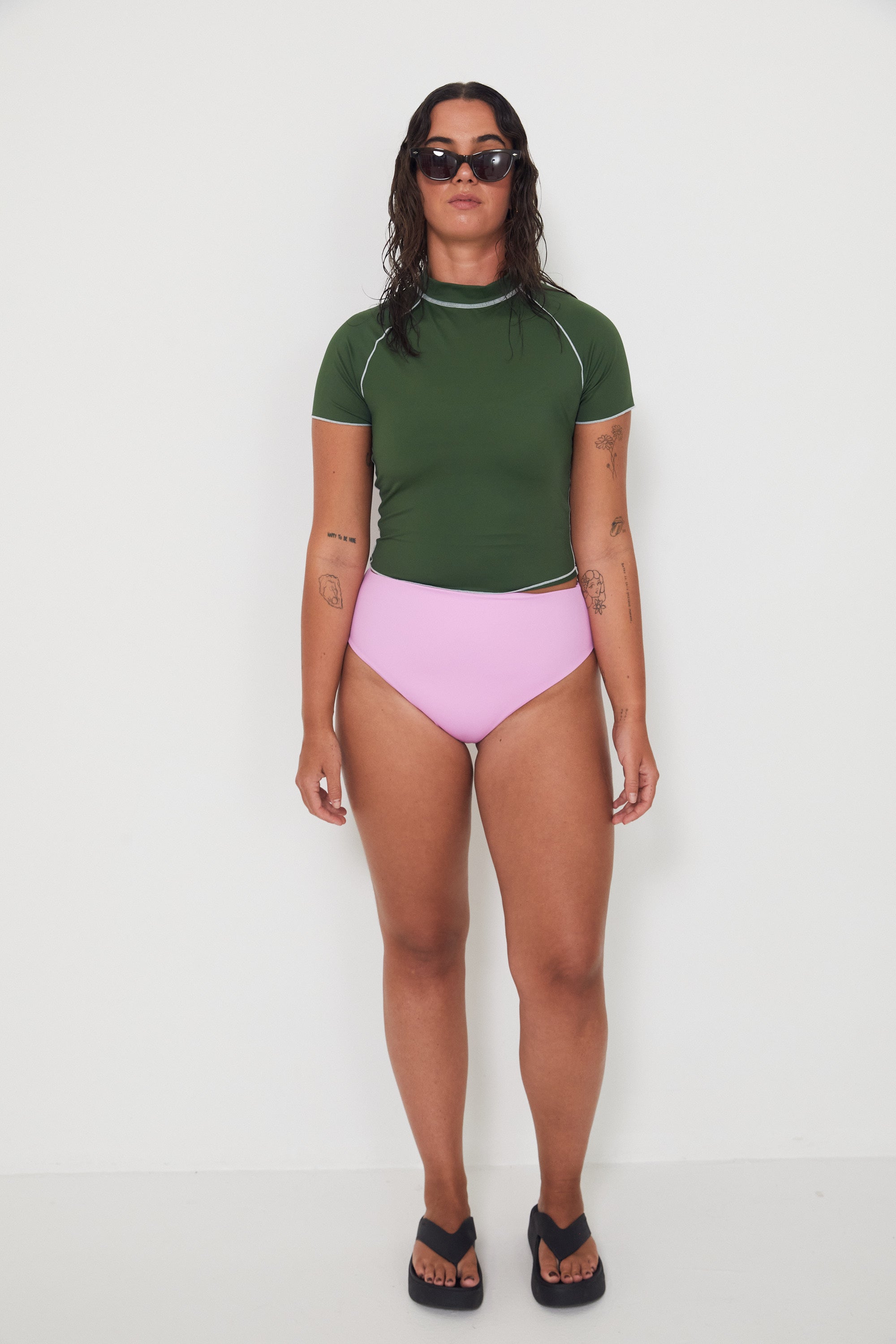 Woman wearing a green olive low rider rashguard and pink drifter swim bottoms with white trim on a plain background